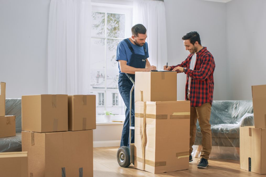 Happy New Homeowner Welcomes Professional Mover with Hand Truck full of Cardboard Boxes, Receives His Goods and Signs on Clipboard