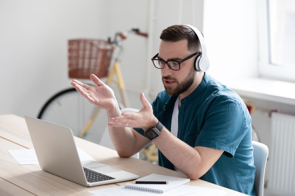 Focused young businessman in eyewear wearing headphones, holding video call with clients on laptop.