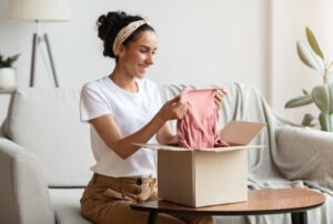 Happy young brunette woman sitting on sofa by wooden table and opening carton box, checking her delivery, holding new clothes and smiling, sarisfied with her order, shopping from home during lockdown