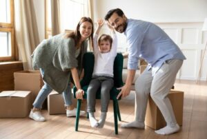 Portrait of excited caucasian young family carry small cute son sit in chair settle in living room in new home, smiling parents have fun unpacking moving relocating to own house with little boy child