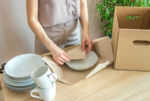 a young girl wraps white porcelain plates in craft paper for sale in an online store. there are empty plates, mugs, and a box on the table