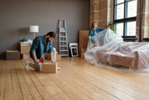 Horizontal long shot of young man and woman getting ready to move to new apartment packing things in boxes and wrapping furniture