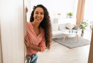 Welcome. Portrait of cheerful woman standing in doorway of modern apartment, greeting visitor and inviting guest to enter her home, happy smiling young lady holding door looking out flat