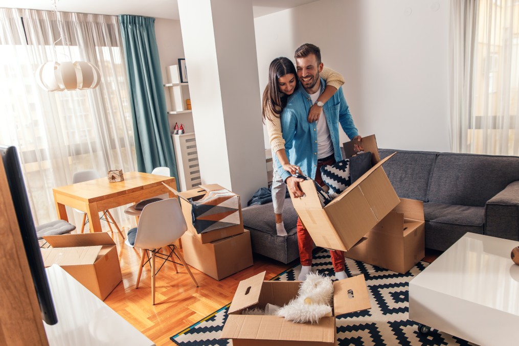 Smiling young couple move into a new home carrying boxes of belongings.