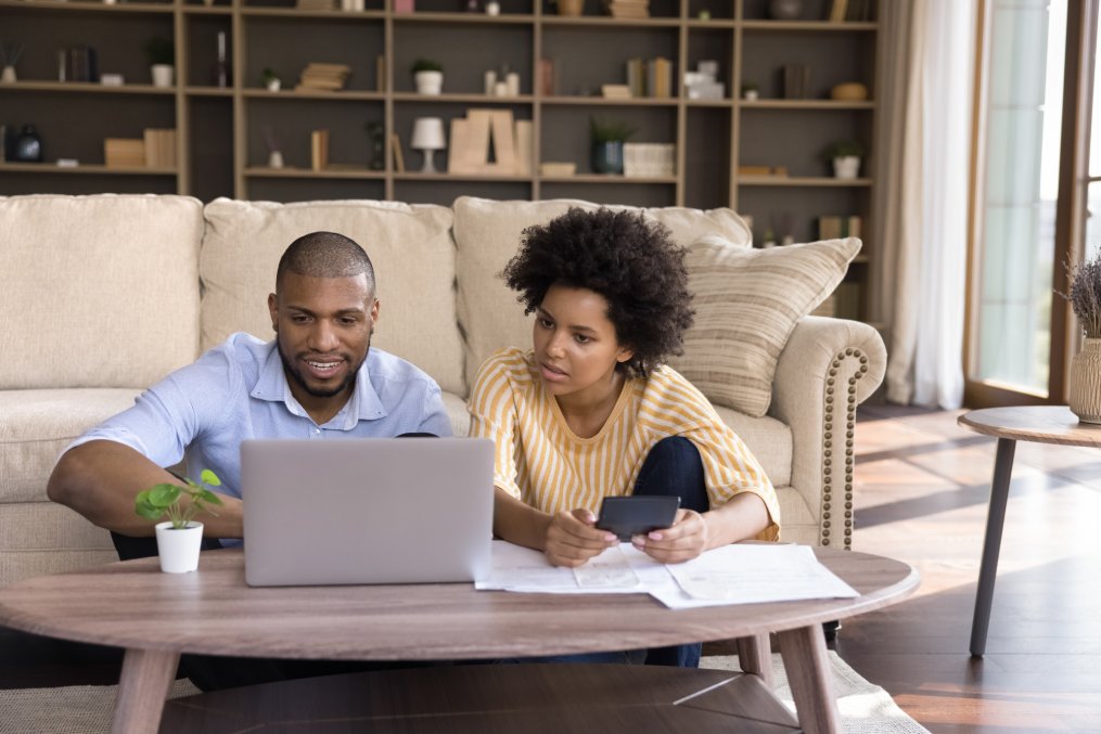 Millennial African American couple using laptop computer at home, making online payments, paying paper bills, insurance, mortgage fees, planning expenses, calculating, money, shopping