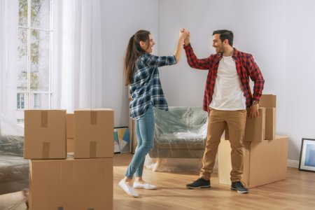 Happy Young Couple Moving in Into New Apartment, Carrying Cardboard Boxes with Stuff, Having Fun, Give High Five. Young Boyfriend and Girlfriend Start Living Together, Unpacking Stuff.