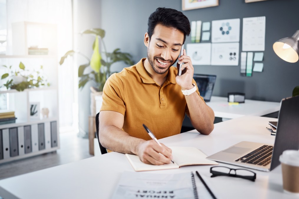 Phone call, happy and Asian man writing notes for reminder, schedule and planning at office desk. Communication, business and male worker on smartphone talking, in discussion and consulting for help