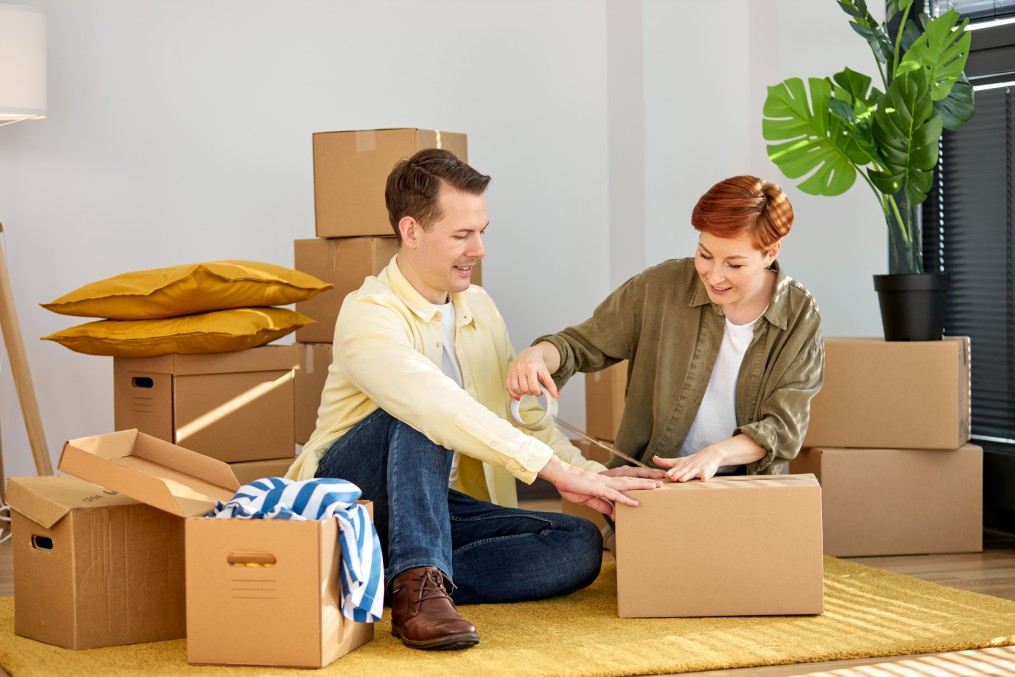 Happy man and woman seal wrap packages boxes with tape dispenser relocate to new house