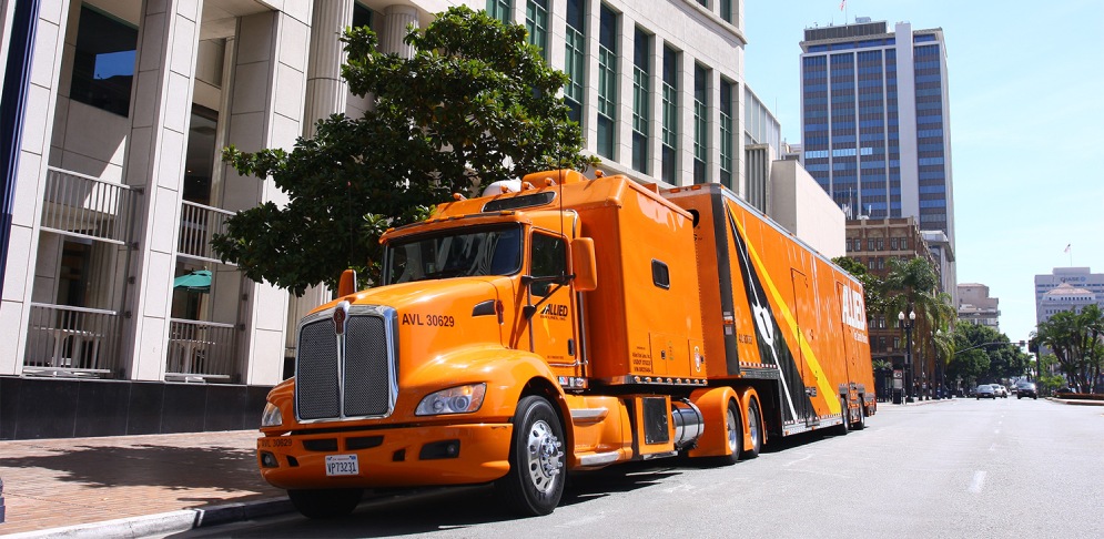 Orange ‘ALLIED’ moving truck on a city street in Orange County, framed by skyscrapers and trees under a clear sky