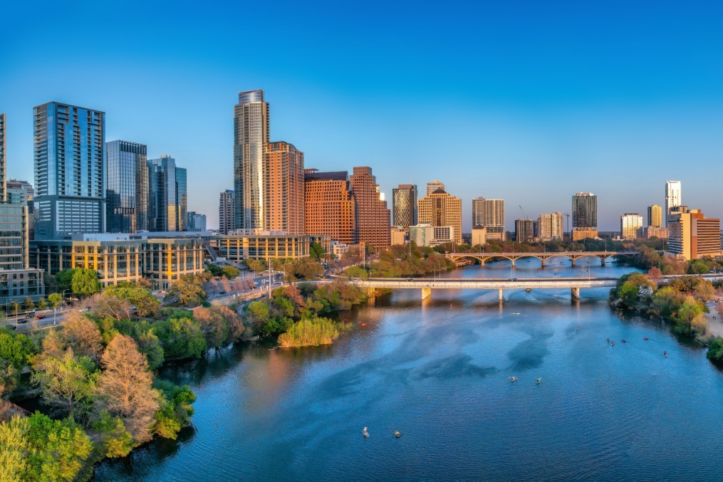 Colorado River near the district urban area of Austin, Texas. There are bridges over the river and a view of skyscraper buildings on the left and large field at the right side against the sunset sky.