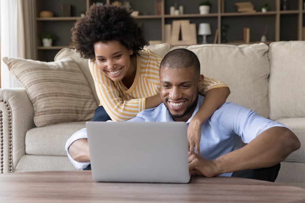 Cheerful African couple look at laptop screen spend carefree weekend at home use modern tech, buying on internet, enjoy distancing communication with family through videocall app. Fun, leisure concept