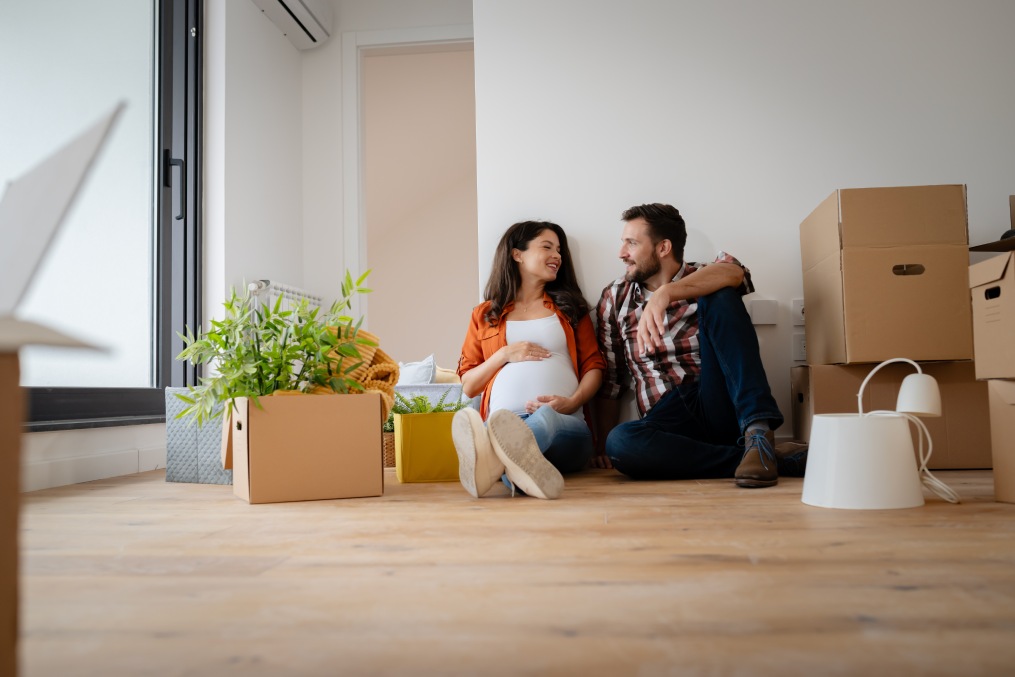 Beautiful young couple expecting a baby just moved into an empty apartment, sitting among cardboard boxes making plans for future. New beginnings