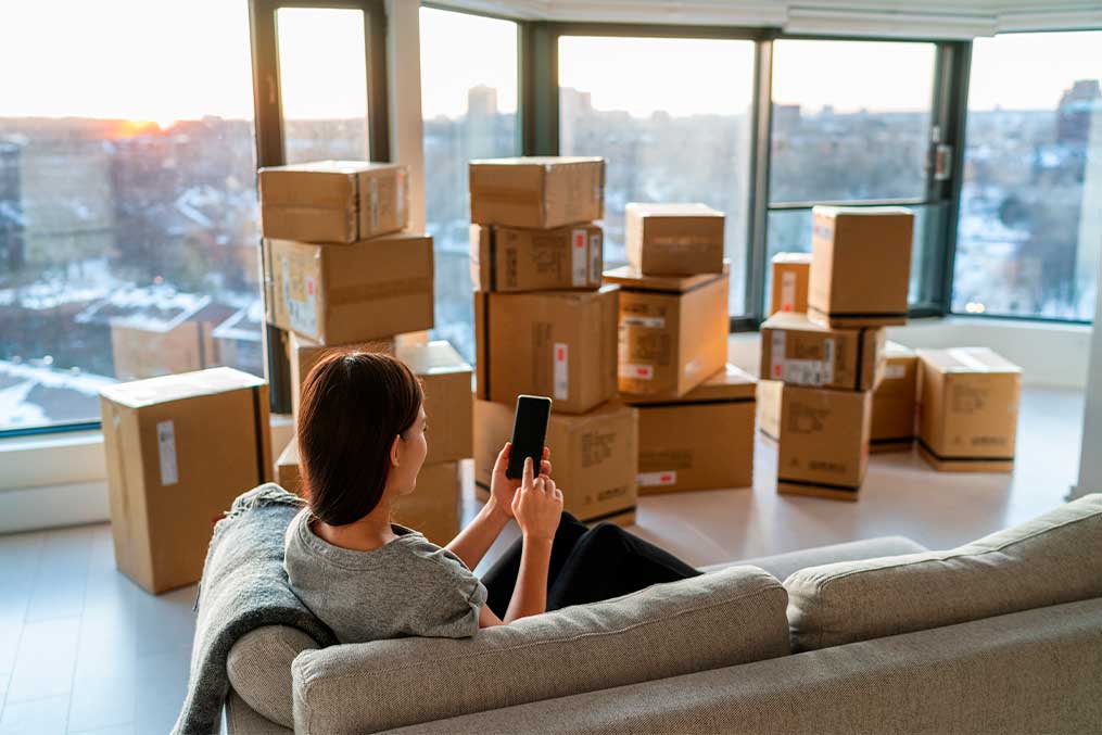 A woman sitting on a couch surrounded by moving boxes, holding a cellphone and appearing to organize her new space.