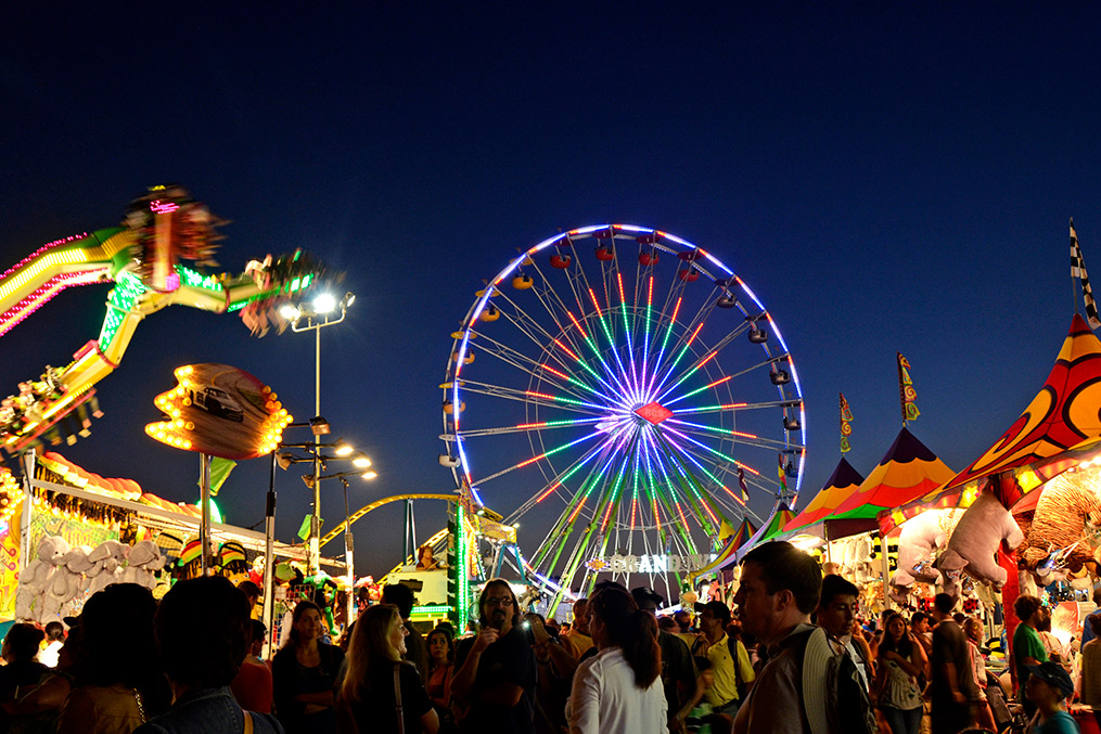 A large crowd enjoys a summer fair with a colorful ferris wheel in the background, highlighting San Diego's vibrant events.