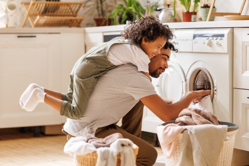 Side view of black child in casual clothes with curly hair smiling and embracing dad loading washing machine during household routine in morning at home