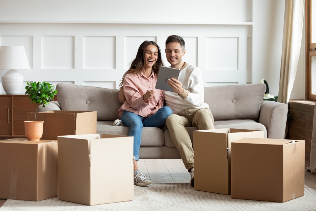 In cozy living room heap of carton boxes, couple sit on sofa with tablet buying furniture on internet, search design ideas, enjoy rest at new home. E-commerce house services, first flat owners concept
