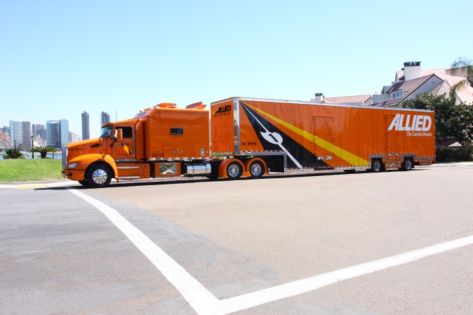 Bright orange Allied moving truck and trailer parked near a city skyline, representing long-distance or cross-country moving services under clear blue skies