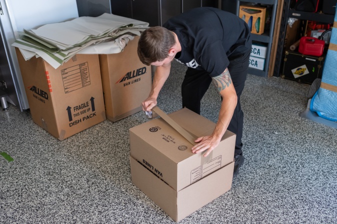 Professional mover sealing a cardboard box with packing tape, surrounded by Allied Van Lines boxes and packing materials, preparing items for a long-distance or cross-country move.