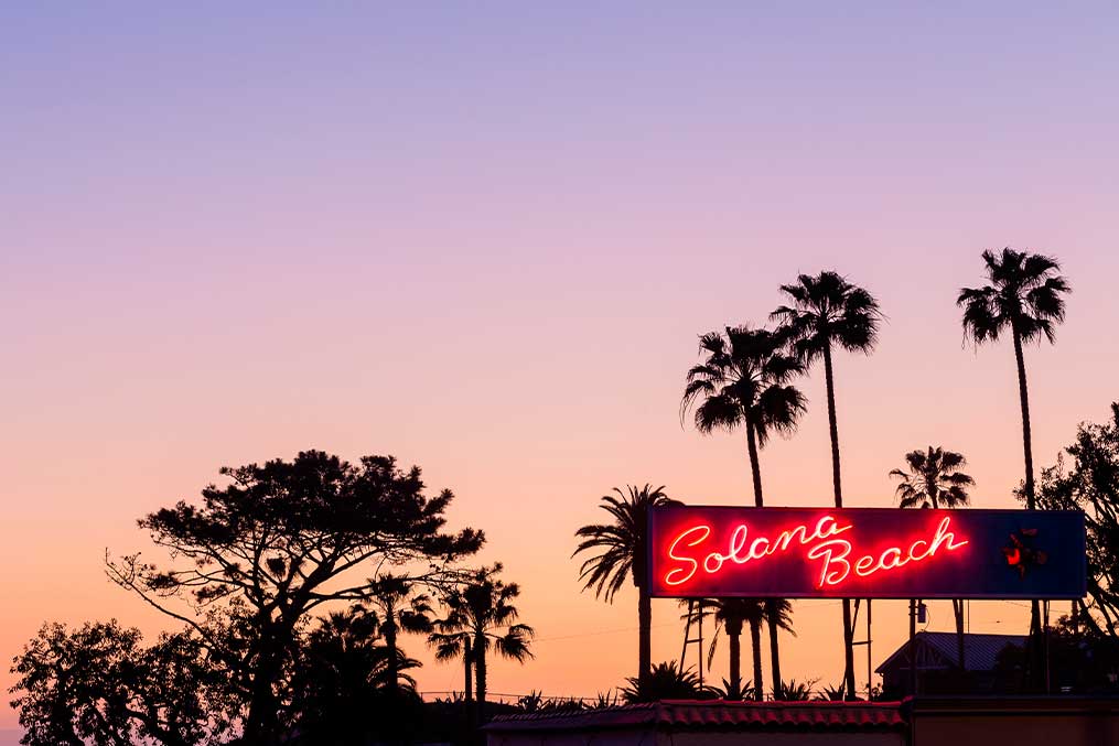 Neon sign with palm trees in the background, promoting "Living in Solana Beach: North County Coastal Guide."