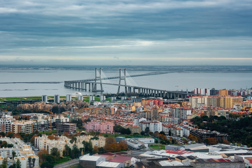 Lisbon, Portugal cityscape faces the Tagus River and Vasco da Gama Bridge. Overcast light softens pastel blocks and silvery water, suggesting a cool quiet day.