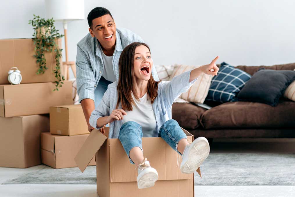 New home, relocation. Happy mixed race young family couple, caucasian girl and latino guy, is playing with a cardboard box, girl sits inside and guy push it, their have fun together at new apartment
