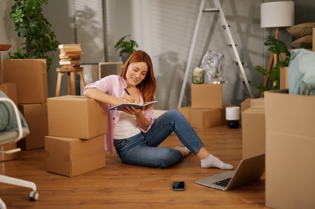 A woman sits on the floor surrounded by cardboard boxes, writing notes in a notebook. She is organizing her thoughts for the move while using a laptop close by.