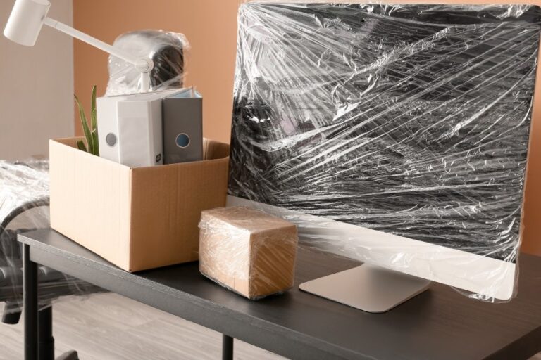 Office desk with computer monitor and cardboard boxes wrapped in plastic for moving or relocation, Transition of a workspace, with personal and professional items packed for transport
